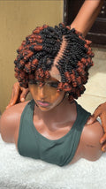 A short, curly wig with black and copper-brown highlights displayed on a mannequin bust. The wig features a center part and voluminous, tightly coiled curls with textured ends.